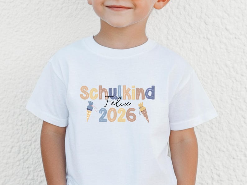 May include: White t-shirt with the text "Schulkind Felix 2026" in colorful letters and decorative ice cream cone graphics. The shirt is made of a soft material and is suitable for children.