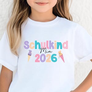 May include: A white t-shirt with the text "Schulkind Mia 2026" in pastel colors. The design includes ice cream cone illustrations. The shirt is likely for a child starting school in 2026.