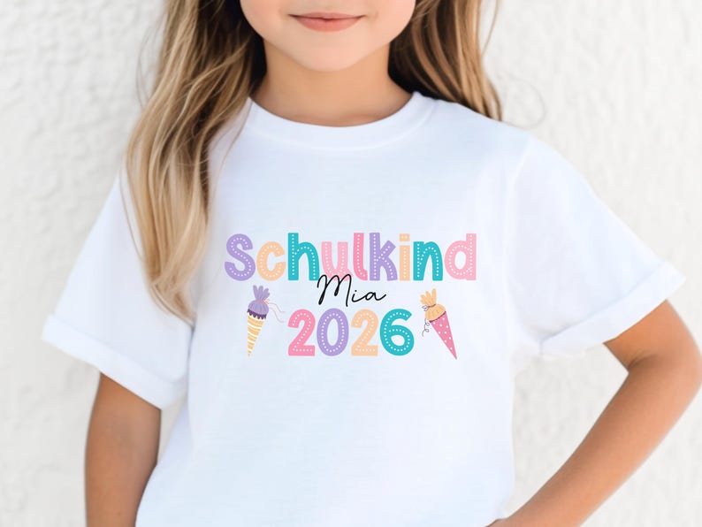 May include: A white t-shirt with the text "Schulkind Mia 2026" in pastel colors. The design includes ice cream cone illustrations. The shirt is likely for a child starting school in 2026.