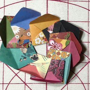 May include: A circular arrangement of small, colorful origami envelopes. The envelopes are made from patterned paper with floral and geometric designs, in shades of blue, green, yellow, and pink. The envelopes are open, revealing their interiors.