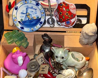 Op de afbeelding: Een houten doos gevuld met diverse snuisterijen, waaronder een Romeo y Julieta sigarenkistdeksel, een miniatuur deegroller, een Solvang windmolenbordje, een metalen eend, een roze eenhoorn badeendje, een keramieken kat, Scrabble-steentjes en diverse metalen lepels.