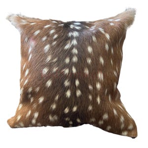 May include: A square throw pillow covered in brown animal hide featuring a pattern of white spots. The texture of the fur is visible across the surface, with a darker brown stripe running vertically down the center of the pillow cover.