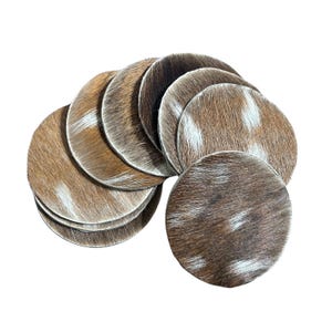 May include: A collection of round coasters with a natural cowhide appearance. The coasters feature a mix of brown, tan, and white patterns. The coasters are arranged in a semi-circular shape, showcasing their texture and design.