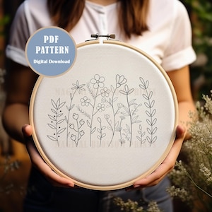 May include: An embroidery hoop with a beige fabric displaying a black line drawing of wildflowers. The text "PDF PATTERN Digital Download" is in a blue circle. The hoop is held by a person. The wooden hoop measures approximately 20 cm in diameter.