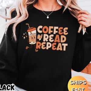 以下が含まれることがあります： 黒色のスウェットシャツ。茶色とオレンジ色の文字で「COFFEE READ REPEAT」と書かれています。本を持った漫画のコーヒーカップも描かれています。スウェットシャツには「BLACK」と「SHIPS FAST」の文字もプリントされています。