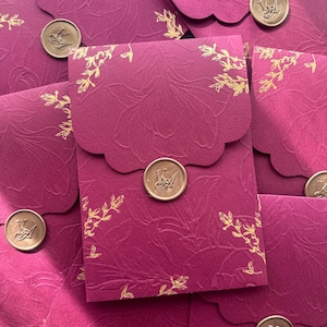 Könnte beinhalten: Magentafarbene Umschläge mit floralem Prägemuster und goldfarbenen Akzenten. Jeder Umschlag ist mit einem goldenen Wachssiegel mit Initialen versehen. Die Umschläge haben eine wellenförmige Klappe.