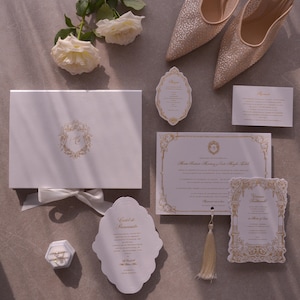 May include: An elegant wedding invitation suite featuring a white box with a gold emblem, matching stationery, and a pair of sparkly heels. The set includes invitation cards, a ring box, and white roses, all arranged on a neutral surface.