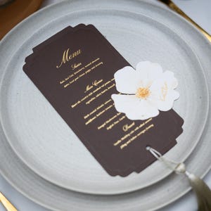 May include: A formal table setting with a dark brown menu card featuring gold text, placed on a gray plate. A white flower with a gold center and the name "Kaitlin" is on the menu. A gold knife and a glass are also visible.
