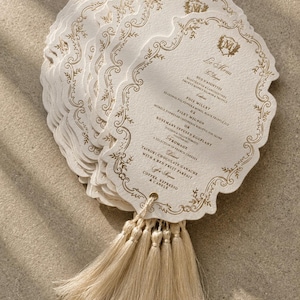May include: A stack of off-white, scalloped-edge menu cards featuring gold foil filigree borders and a central monogram. Each card includes printed text detailing a multi-course meal and is finished with a beige tassel attached through a metal eyelet.