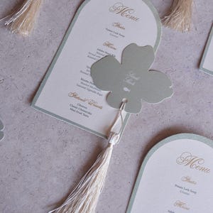 Könnte beinhalten: Elegante Hochzeitsmenükarten mit einem hellgrünen Rand und goldener Schrift. Jede Karte zeigt ein Menü mit Vorspeisen, Hauptgerichten und Desserts. Ein kleeblattförmiges Etikett mit Quaste sorgt für eine dekorative Note.