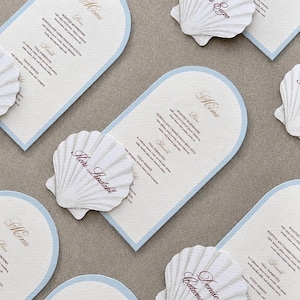 May include: Arch-shaped paper menus with light blue borders and white textured cardstock. Each menu features a white, three-dimensional seashell cutout attached to the front, displaying a guest name in brown script and a printed food list.