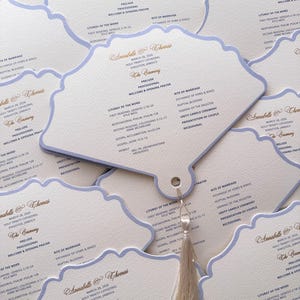 May include: Off-white wedding ceremony programs with a pale blue border and gold text. Each program has a unique scalloped shape and is tied with a cream-coloured tassel. The programs list the ceremony details.