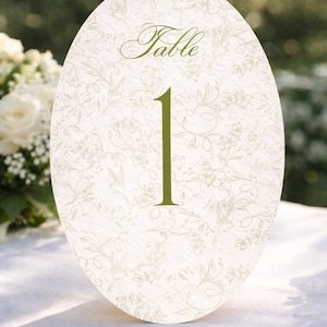 Könnte beinhalten: Ovale Tischnummerkarte mit floralem Muster und dem Wort "Table" in Schrift. Die Nummer "1" ist in einem Grünton gehalten. Die Karte befindet sich auf einer weißen Tischdecke mit verschwommenem Hintergrund aus Grün.
