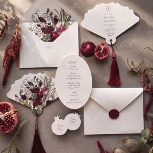 May include: An assortment of wedding stationery featuring white envelopes, fan-shaped cards, and oval cards. The designs include illustrations of pomegranates and floral elements in burgundy and green. A red tassel and a pomegranate half are also visible.
