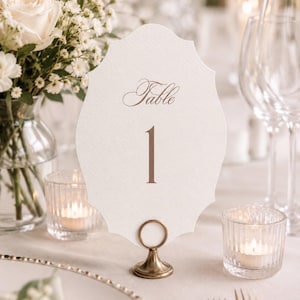 May include: A white scalloped table number card featuring the word Table in elegant script above the number 1 in a serif font. The card is held upright by a small, circular, metallic gold stand with a flared base, positioned on a table.