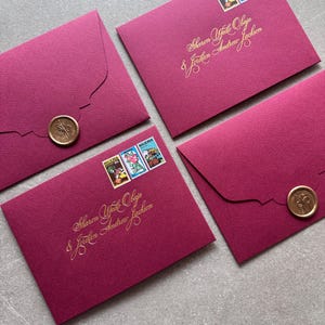 Puede incluir: Cuatro sobres magenta con letras doradas y sellos. Dos sobres tienen sellos que dicen "NIGERIA". La escritura dice "Sharon Yfode Obaje & Jordan Andrew Jackson". Dos sobres están sellados con un sello de cera dorado.