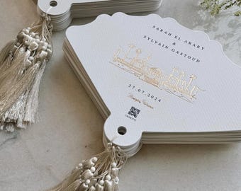 May include: Fan-shaped wedding programs made of white cardstock with scalloped edges and beige tassels. One side features a gold foil illustration of a building with palm trees, the names Sarah El Araby and Sylvain Gastoud, the date 27.07.2024, and a QR code.