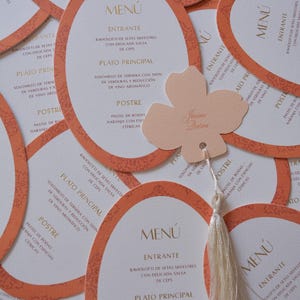 Peut inclure: Cartes de menu de mariage avec un design ovale et une bordure orange. Chaque carte présente les détails du menu en texte doré. Une étiquette en forme de fleur pêche avec un gland est suspendue aux cartes.