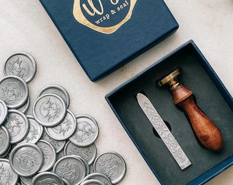 May include: A set of silver wax seals with a floral design and the initials "M & M" in the center. The seals are in a blue box with a gold logo that says "WS Wrap & Seal". There is also a silver wax seal stamp and a wooden handle in a separate blue box.