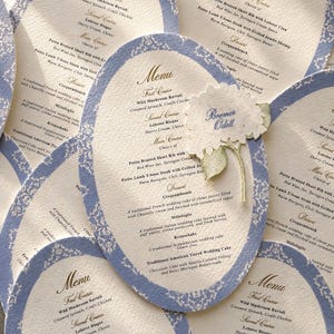 May include: An assortment of elegant wedding menu cards. Each card is oval-shaped with a light blue border and intricate white floral patterns. The menu features multiple courses, including Wild Mushroom Ravioli and Lobster Bisque. A floral embellishment with the name "Boomer Odell" is also present.