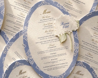 Könnte beinhalten: Eine Auswahl an eleganten Hochzeitsmenükarten. Jede Karte hat eine ovale Form mit einem hellblauen Rand und aufwendigen weißen Blumenmustern. Das Menü bietet mehrere Gänge, darunter Wildpilz-Ravioli und Hummer-Bisque. Ein Blumenschmuck mit dem Namen "Boomer Odell" ist ebenfalls vorhanden.