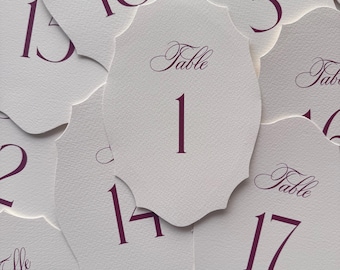 May include: A collection of off-white, scalloped-edge table number cards featuring dark purple text. Each card displays the word Table in a cursive script above a large numeral, including numbers 1, 3, 13, 14, 17, and 18, printed on textured paper.