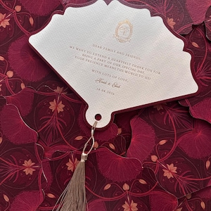 May include: A burgundy and cream wedding invitation in a fan shape, featuring a floral design. The cream card has gold text and a tassel. The text reads "Dear family and friends, we want to extend a heartfelt thank you..." and the date 18.04.2026.