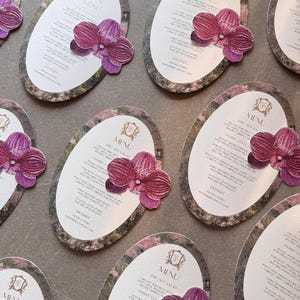 May include: Oval menu cards with floral borders and a purple orchid accent. Each card displays the word "MENU" at the top, with a list of dishes below. The cards are arranged on a grey surface.