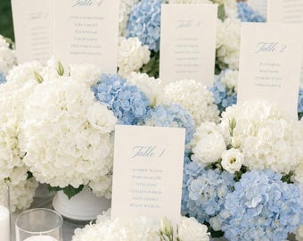 Könnte beinhalten: Mehrere Blumenarrangements aus weißen und hellblauen Hortensien, die als Tischdekoration dienen. Jedes Arrangement enthält eine vertikale cremefarbene Karte mit blauem Text, der die Tische eins bis vier identifiziert und die Namen der Gäste auflistet.