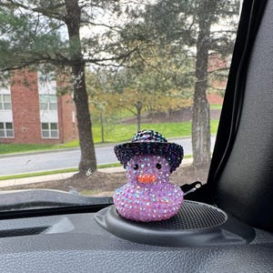 May include: A purple and black jeweled rubber duck wearing a black cowboy hat with a purple band. The duck is sitting on a black car dashboard.