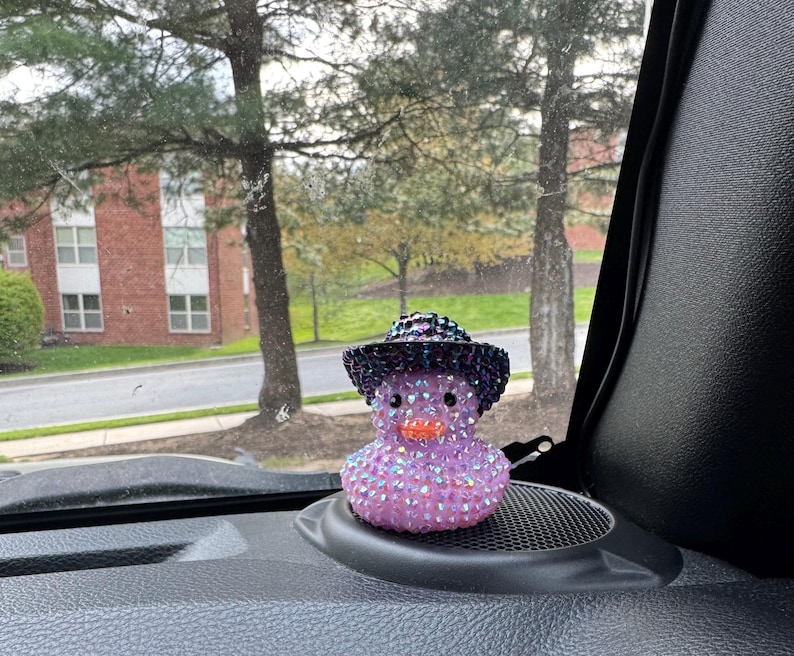 May include: A purple and black jeweled rubber duck wearing a black cowboy hat with a purple band. The duck is sitting on a black car dashboard.