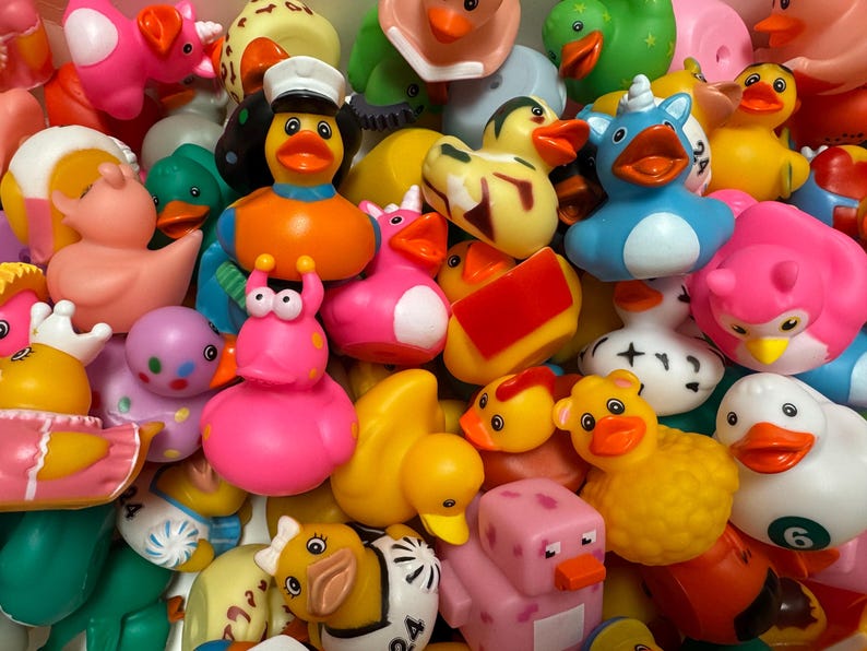 May include: A collection of colorful rubber duck toys in various designs. The ducks are in shades of yellow, pink, blue, and green, with some wearing hats or having unique features like horns or crowns. The toys are a mix of sizes and shapes.