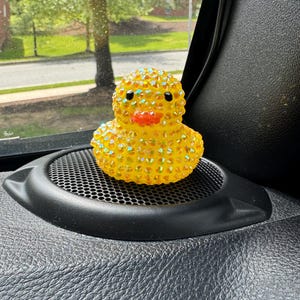 May include: A yellow rubber duck covered in iridescent rhinestones. The duck has black eyes and an orange beak. It is sitting on a black speaker grill in a car. The background shows a street and trees.