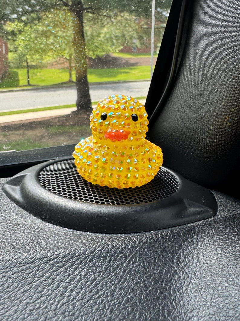 May include: A yellow rubber duck covered in iridescent rhinestones. The duck has black eyes and an orange beak. It is sitting on a black speaker grill in a car. The background shows a street and trees.