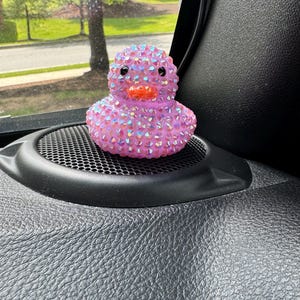 May include: A pink rubber duck covered in iridescent rhinestones. The duck has black eyes and an orange beak. It is sitting on a black speaker grill.