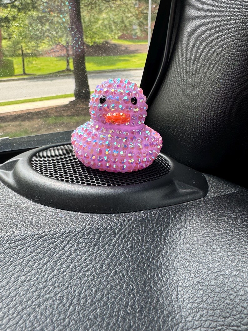 May include: A pink rubber duck covered in iridescent rhinestones. The duck has black eyes and an orange beak. It is sitting on a black speaker grill.