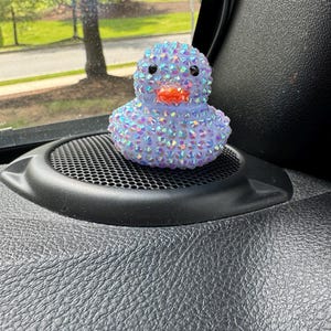 May include: A bedazzled rubber duck figurine. The duck is covered in iridescent rhinestones, giving it a sparkling appearance. It has black eyes and an orange beak. The duck is sitting on a black speaker.