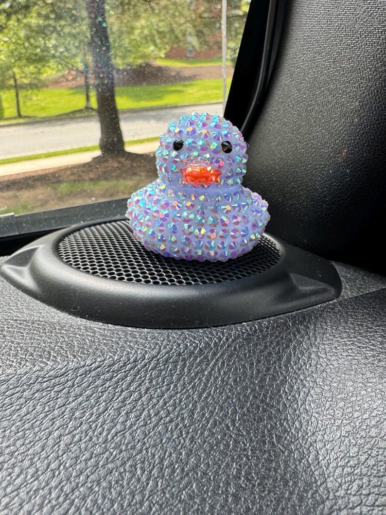 May include: A bedazzled rubber duck figurine. The duck is covered in iridescent rhinestones, giving it a sparkling appearance. It has black eyes and an orange beak. The duck is sitting on a black speaker.