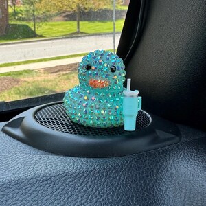 May include: A teal-colored duck figurine covered in iridescent rhinestones, with a light blue cup and straw. The duck has black eyes and an orange beak. The figurine is sitting on a black surface.