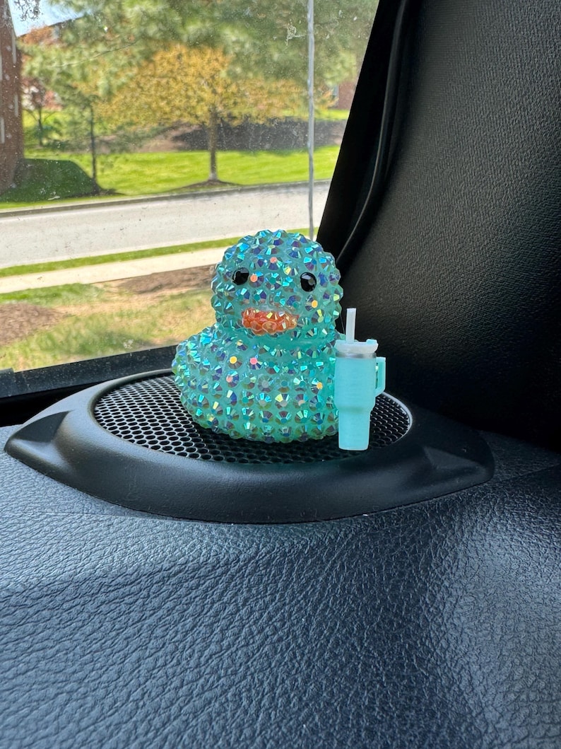 May include: A teal-colored duck figurine covered in iridescent rhinestones, with a light blue cup and straw. The duck has black eyes and an orange beak. The figurine is sitting on a black surface.