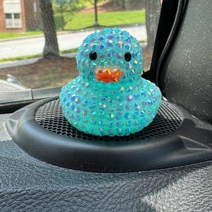May include: A turquoise duck figurine covered in iridescent rhinestones. The duck has black eyes and an orange beak. It is sitting on a black speaker grill.