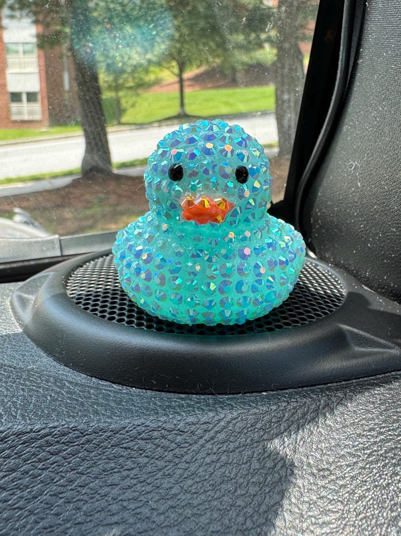 May include: A turquoise duck figurine covered in iridescent rhinestones. The duck has black eyes and an orange beak. It is sitting on a black speaker grill.