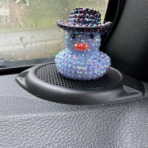 May include: A blue and iridescent, rhinestone-covered rubber duck wearing a black and purple rhinestone cowboy hat. The duck is sitting on a black car dashboard.