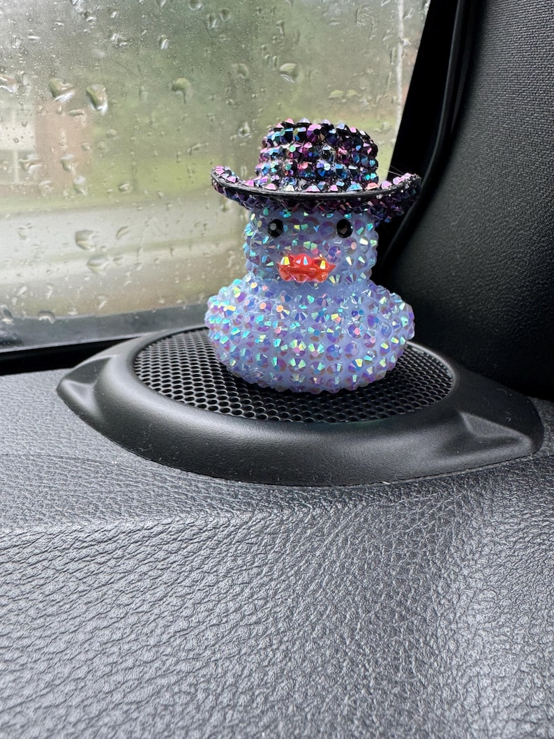May include: A blue and iridescent, rhinestone-covered rubber duck wearing a black and purple rhinestone cowboy hat. The duck is sitting on a black car dashboard.