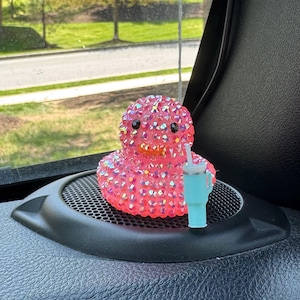 Könnte beinhalten: Eine pinkfarbene, mit Strass besetzte Entenfigur mit schwarzen Augen und einem orangefarbenen Schnabel. Daneben befindet sich ein kleiner, hellblauer Becher mit einem weißen Strohhalm. Die Gegenstände stehen auf einer schwarzen Lautsprecherabdeckung, mit einem Fenster und Grün im Hintergrund.