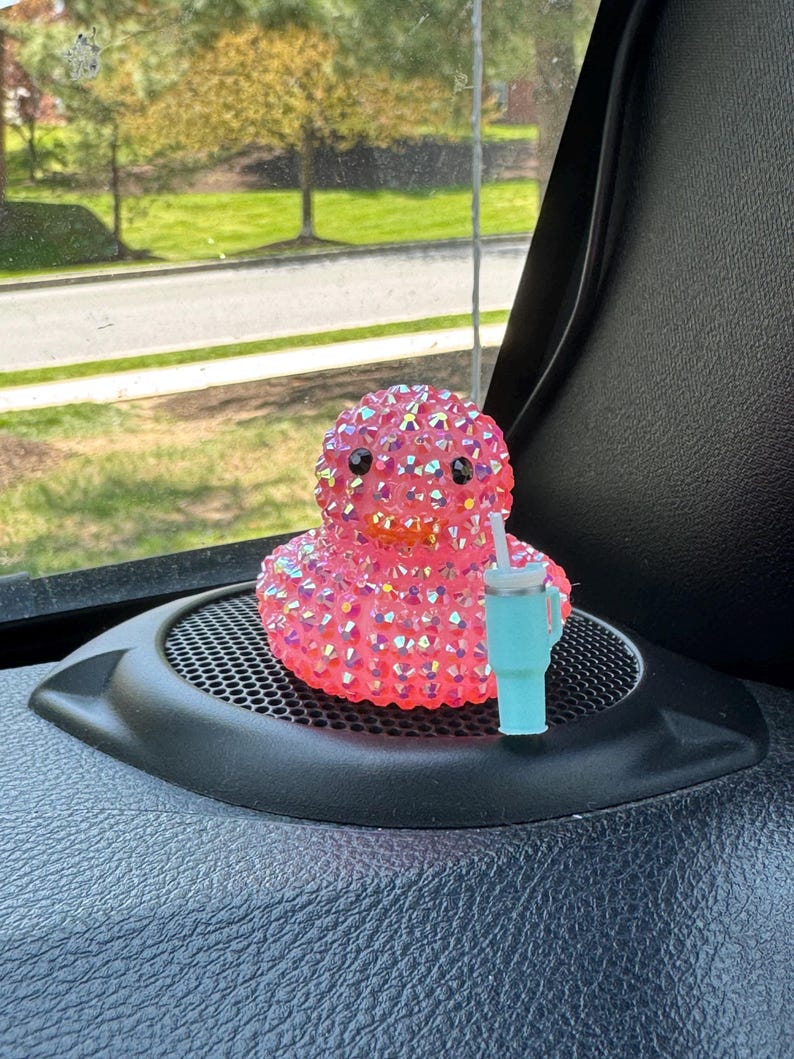 May include: A pink, bedazzled duck figurine with black eyes and an orange beak. The duck is next to a small, light blue tumbler with a white straw. The items are on a black speaker cover, with a window and greenery in the background.