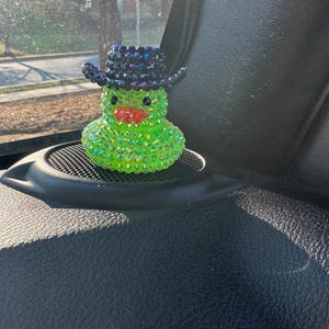 May include: A green rubber duck with a black cowboy hat covered in rhinestones. The duck is hanging from a rearview mirror in a car.