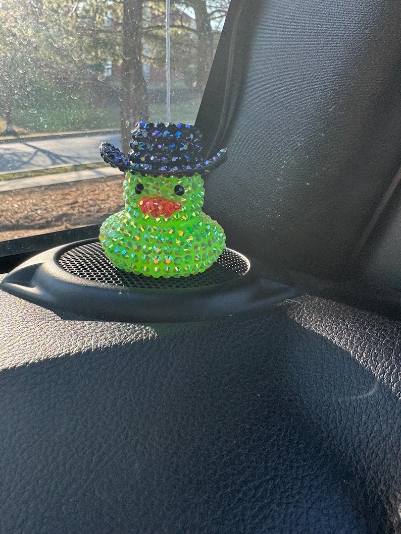 May include: A green rubber duck with a black cowboy hat covered in rhinestones. The duck is hanging from a rearview mirror in a car.