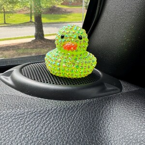 May include: A bedazzled, lime green rubber duck with a bright orange beak and black eyes. The duck is covered in iridescent rhinestones, giving it a sparkling appearance. It sits on a black speaker grill, with a blurred outdoor scene in the background.