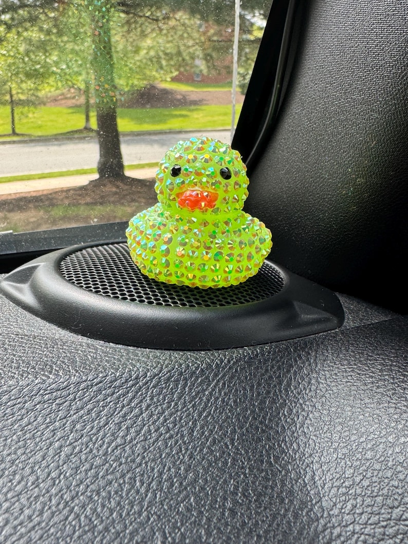 May include: A bedazzled, lime green rubber duck with a bright orange beak and black eyes. The duck is covered in iridescent rhinestones, giving it a sparkling appearance. It sits on a black speaker grill, with a blurred outdoor scene in the background.